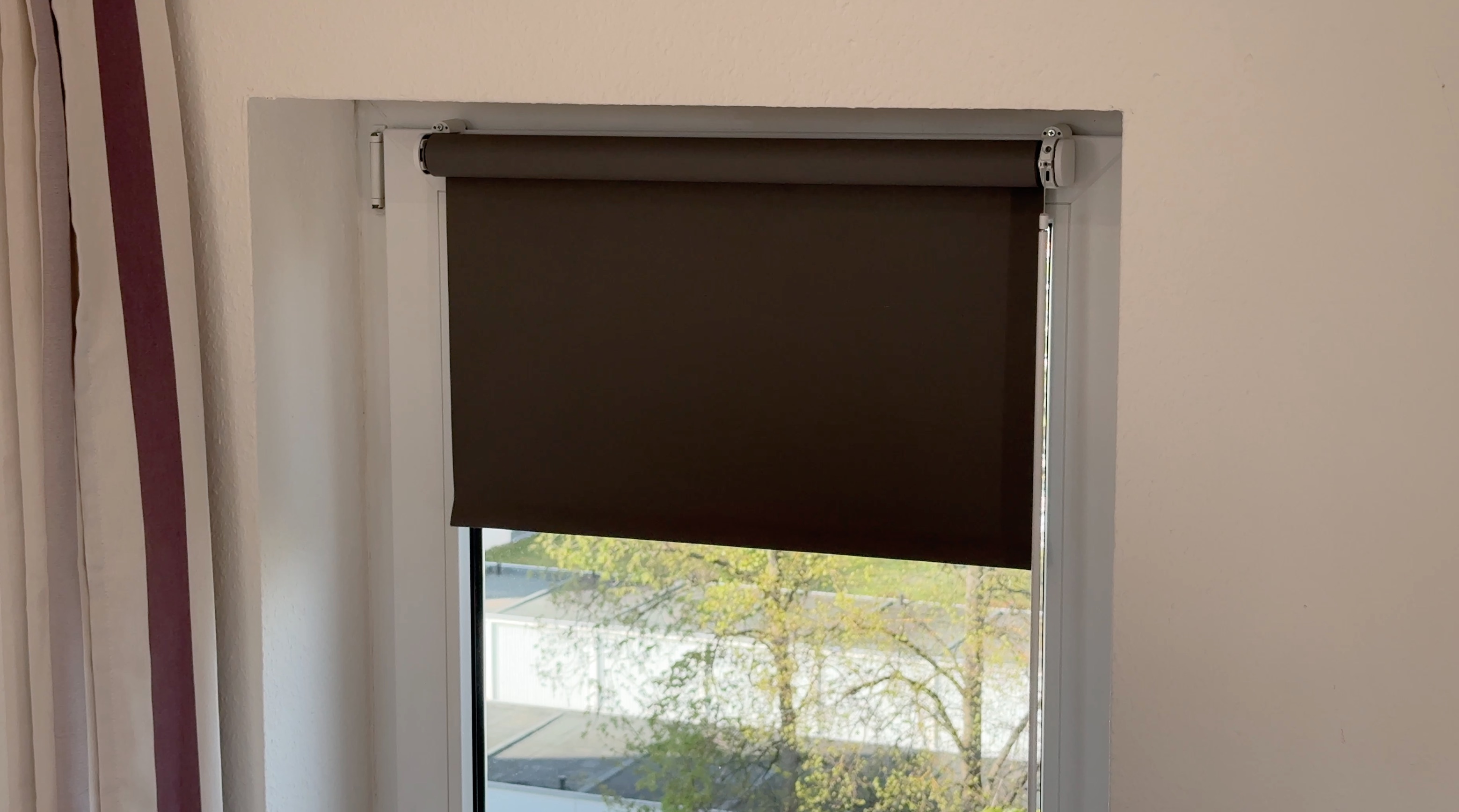 Partially lowered dark roller blind on a window with trees outside